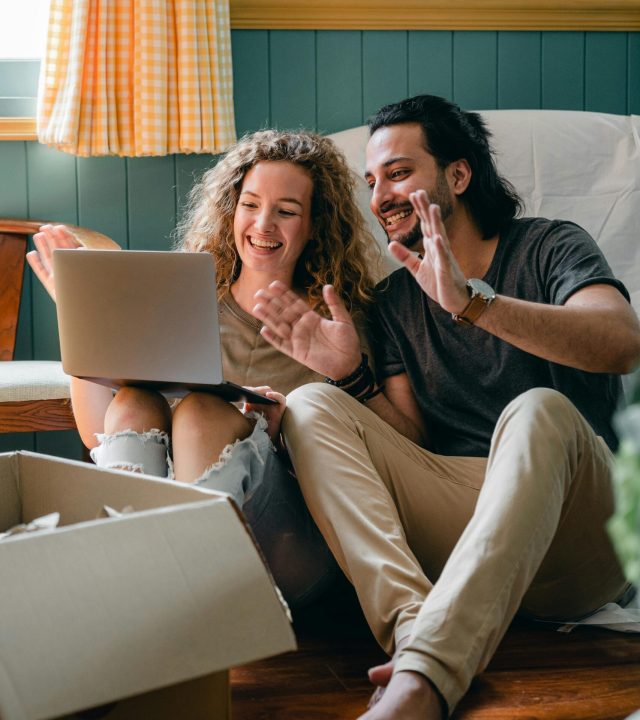 Full body happy smiling diverse couple in casual wear sitting on floor near cardboard boxes in new apartment and chatting via netbook